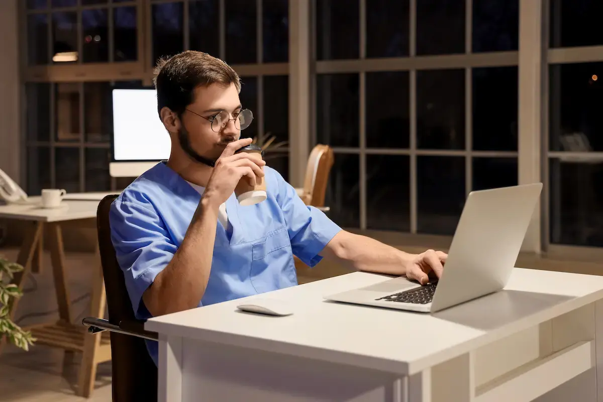 Nurse working at a clinic overnight, drinking coffee and working on a laptop