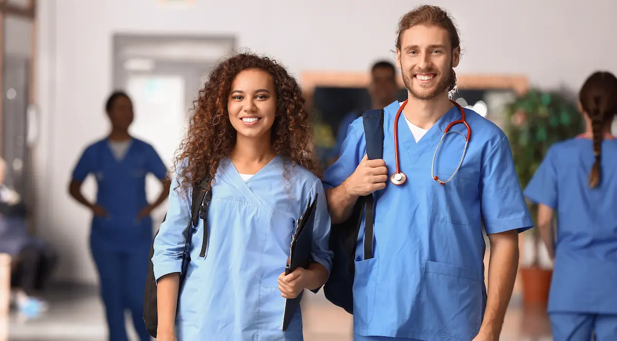 Two nursing students standing in a hallway together smiling and holding backpacks